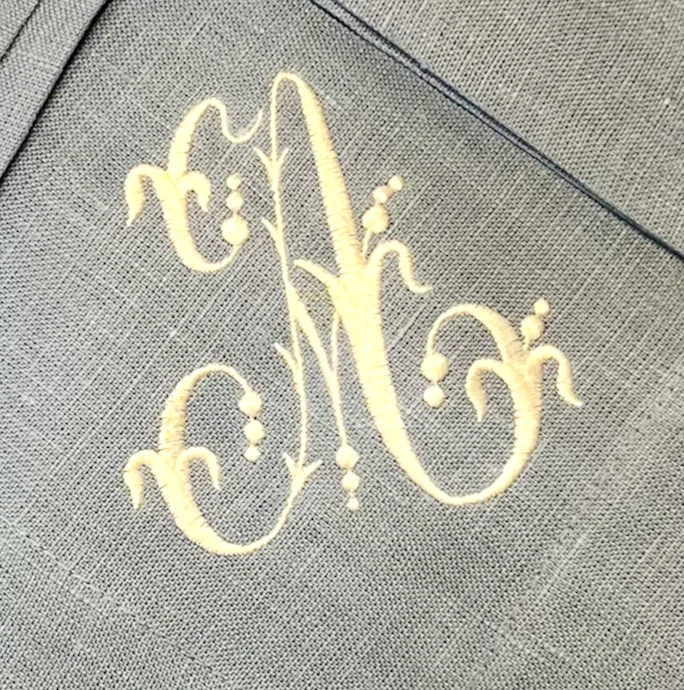 A swirling embroidered monogram letter A in cream thread on grey linen.