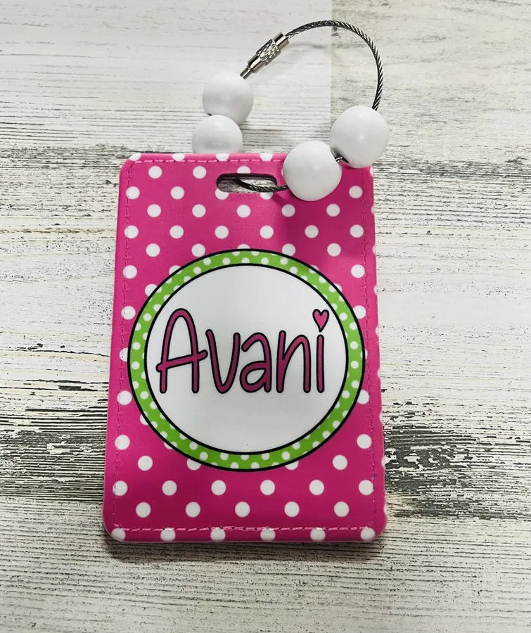 A pink polka-dot bag tag she made, the name Avani printed inside a green spotted circle, strung on white wooden beads.